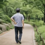공원 산책로에서 허리 옆쪽에 손을 얹고 천천히 걷는 중년 남성의 뒷모습