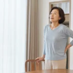 집 창가에서 잠시 멈춰 숨을 고르며 몸 상태를 살피는 중년 한국인