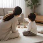 거실 매트에 앉은 세 돌 무렵 아이가 장난감을 가리키고 마주 앉은 보호자가 아이 얼굴을 바라보며 말을 건네는 장면