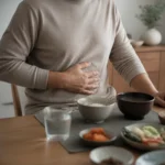 속이 더부룩한 중년 남성이 식탁 앞에서 배를 손으로 누르며 불편한 표정을 짓는 모습