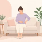 여성이 소파에 앉아 아랫배와 골반 옆쪽을 손으로 짚고 있는 모습