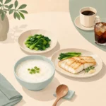 식탁 위에 담백한 죽과 데친 채소, 구운 흰살생선이 놓여 있고 옆에 커피잔과 탄산음료가 밀려나 있는 장면